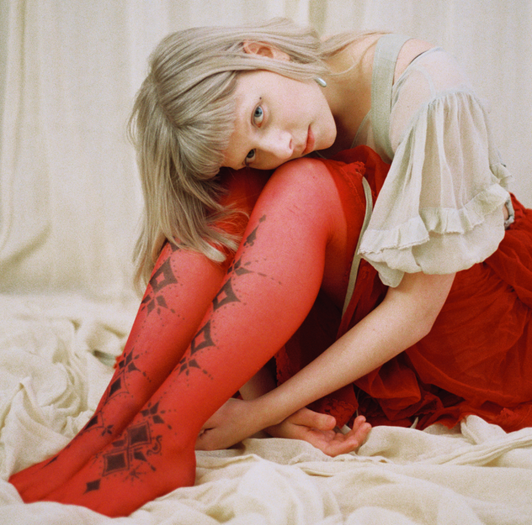 Artisten aurora sitter og omfavner beina sine på en myk hvit tøybakgrunn, ikledd røde strømpebukser og en hvit kjole, hun kikker i kamera