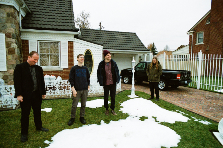 Bandmedlemmene i Protomartyr står foran et hus med en lastebil parkert foran, de er på en plen som er delvis dekket med snø, og ser blide ut.