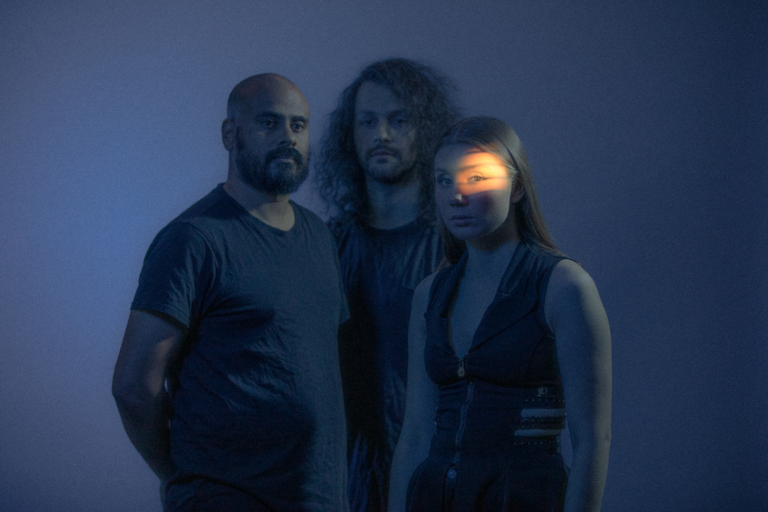 Bildet viser de tre bandmedlemmene i Isák stående i mørket, et lite lyspunkt treffer ansiktet til Ella Marie Hætta Isaksen